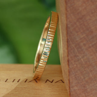 A close-up of a slim gold wedding ring with a hammered tree bark texture, placed on a wooden surface.