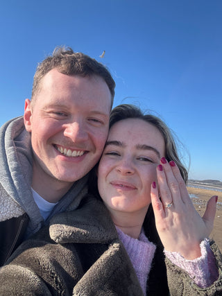 A couple who got engaged with a Lilia Nash handmade sapphire engagement ring. Seaside background.