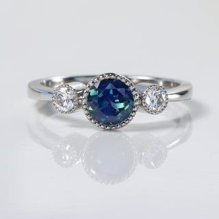 Close-up of platinum Parti sapphire and diamond trilogy ring on reflective background.
