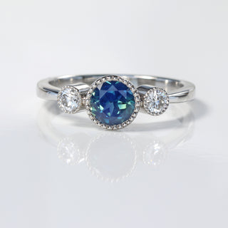 Handmade platinum Parti sapphire and diamond trilogy engagement ring on reflective surface.