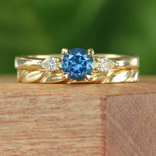 Blue sapphire trilogy engagement ring with pear diamond accents and matching gold leaves wedding band, on a wooden box with green background.