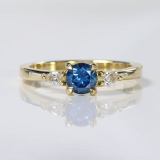 Handmade 18ct gold blue sapphire trilogy engagement ring with pear cut diamonds on reflective surface.