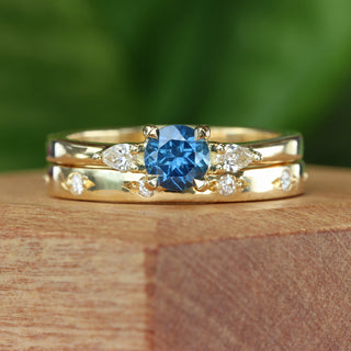 Blue sapphire trilogy engagement ring with pear diamond accents and a matching diamond set wedding band, on a wooden box with green background.