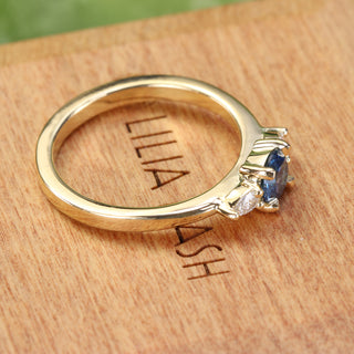 Blue sapphire and diamond trilogy engagement ring photographed at an angle on wood.