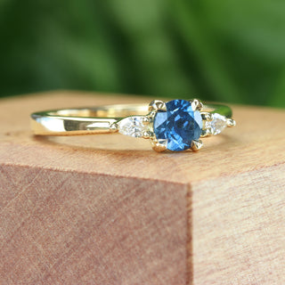 Side angle of fairtrade blue sapphire and diamond trilogy engagement ring on a wooden box with green backdrop.