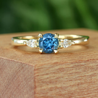 Handmade 18ct gold trilogy engagement ring with blue sapphire and pear diamonds on wooden surface.