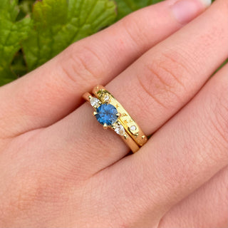blue sapphire and diamond trilogy band with diamond moon and stars wedding band, shown in the hand with green foliage background.
