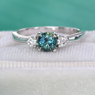 Close-up of teal sapphire and diamond trilogy engagement ring in handmade platinum on green background.