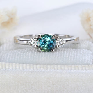 Platinum trilogy engagement ring with teal sapphire and diamonds photographed on pale fabric.