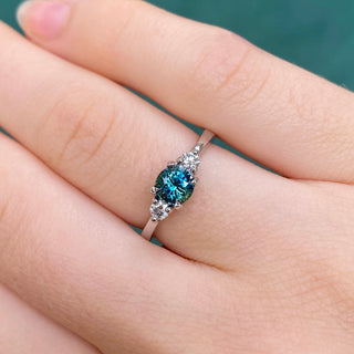 Platinum teal sapphire trilogy engagement ring with diamond accents shown on hand.