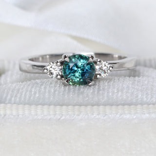 Close-up of handmade platinum trilogy engagement ring featuring teal sapphire and round diamonds.