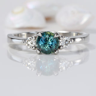 Platinum trilogy engagement ring with teal sapphire and two round diamonds on reflective surface.