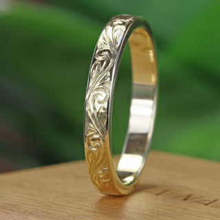 A 3mm wide 18ct gold wedding band with a flat profile, featuring a floral scroll pattern engraved around the outer band shown standing vertically on a wooden box