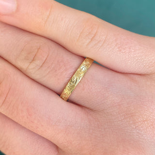 A 3mm wide 18ct gold wedding band with a flat profile, featuring a floral scroll pattern engraved around the outer band shown on a finger