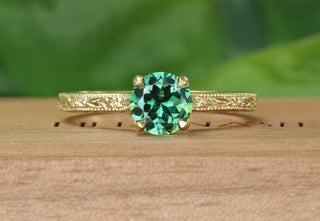 Handmade sustainable 18ct gold green sapphire solitaire engagement ring with hand engraved orange blossom and leaves pattern. Resting on a wooden box with green background