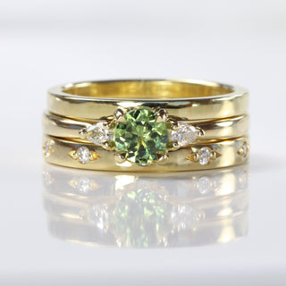 Handmade Stacking ring set of a green sapphire and diamond trilogy ring, 2mm flat profile wedding band and diamond set eternity ring, on a reflective grey background.