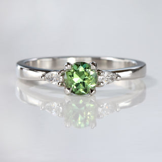Handmade platinum trilogy engagement ring with green sapphire and pear cut diamonds on reflective surface.