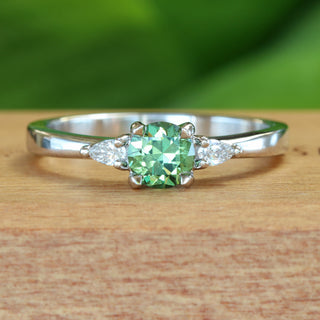 Handmade platinum trilogy engagement ring with green sapphire and pear cut diamonds on wooden ring box.