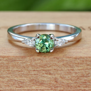 Platinum green sapphire and diamond trilogy engagement ring photographed on wooden background.