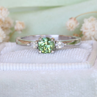 Close-up of platinum trilogy engagement ring with green sapphire and pear diamonds on green background.