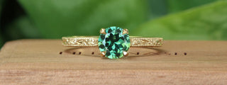 Banner of a 18ct gold green sapphire solitaire engagement ring with hand engraved floral detail