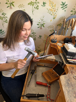 Lilia Nash holding a jewellery making hammer, making a handmade wedding ring at her workshop desk.