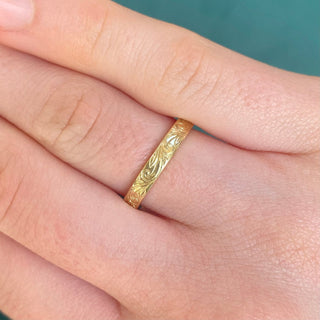 A 3mm wide 18ct gold wedding band with a flat profile, featuring a floral scroll pattern engraved around the outer band shown on a finger