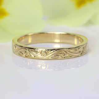18ct gold floral scroll engraved ring on a white surface with green leaves in the background