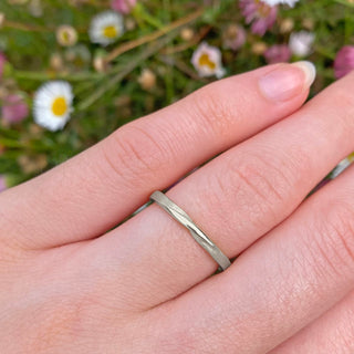 Slim handmade platinum wedding ring with a twist at the front of the band. Lilia Nash's signature spun silk finish is applied to the 2mm wide band. Shown being worn on the  and with daisy background