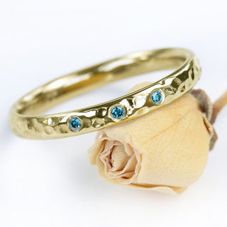 9ct gold hammered wedding band with three blue diamonds set at the front. Displayed resting on a yellow rose on a white background