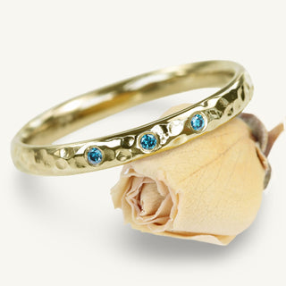 9ct gold hammered wedding band with three blue diamonds set at the front. Displayed resting on a yellow rose on a white background