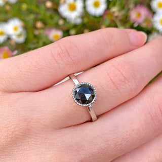 Handmade platinum solitaire ring set with a large 1.9 carat rose cut natural black diamond. The setting has milgrain detail around the stone. Photographed being worn on the hand with daisy backdrop