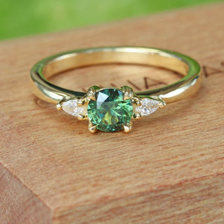 Close up of bespoke 18ct gold dark green sapphire and diamond trilogy engagement ring on a branded wooden box