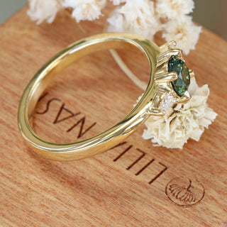 Top view of Bespoke 18ct gold dark green sapphire and diamond trilogy engagement ring on a branded wooden box with white flowers