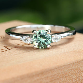 Bespoke platinum mint green sapphire and diamond trilogy engagement ring on a branded wooden box with green background