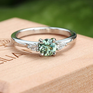 Top view of Bespoke platinum mint green sapphire and diamond trilogy engagement ring on a branded wooden box with green background