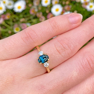 Sustainable dark teal sapphire and diamond trilogy engagement ring, handmade from solid 18ct yellow gold. Shown being worn on the hand, with daisy backdrop