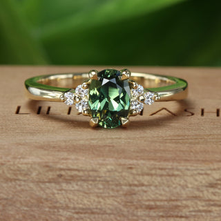 18ct gold cluster engagement ring with an oval green sapphire and side diamonds on a branded wooden box and blurred green background