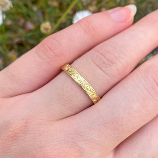 Sustainable 18ct gold 3mm wide wedding ring with a hand engraved floral scroll pattern around the band. Displayed being worn on the hand with daisies in the background
