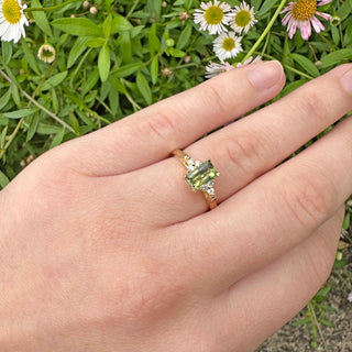 18ct gold Emerald cut Green sapphire and diamond cluster engagement ring being worn on the hand with daisy background