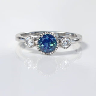Handmade platinum Parti sapphire and diamond trilogy engagement ring on reflective surface.