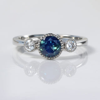 Close-up of platinum Parti sapphire and diamond trilogy ring on reflective background.