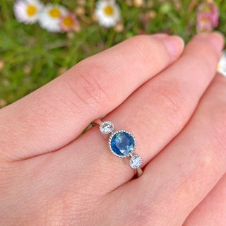 Handmade 950 platinum trilogy engagement ring bezel set with a fair trade dark ocean teal Parti sapphire and natural white diamonds either side. The settings have pretty milgrain edging. Photographed worn on the finger with daisies