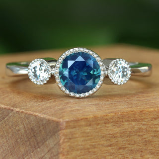 Handmade platinum Parti sapphire and diamond trilogy engagement ring on wooden surface.