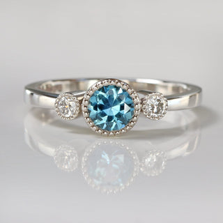 Handmade platinum trilogy engagement ring with teal blue Montana sapphire and Canadian diamonds on reflective surface.