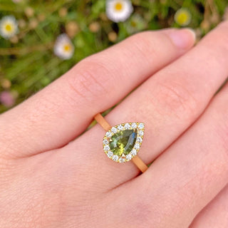 Kynance 18ct gold engagement ring set with a fair trade pear cut green sapphire with a halo of natural white diamonds. Displayed on the hand with daisies