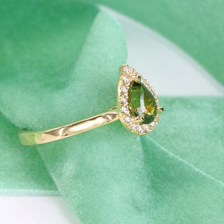 Pear cut green sapphire and diamond halo engagement ring shown at an angle on green fabric.