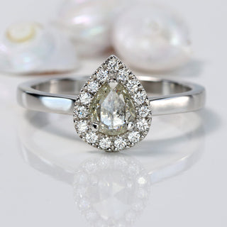 Handmade platinum pear diamond halo engagement ring with pavé setting on reflective surface.