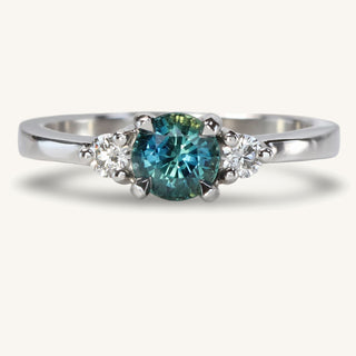 Platinum trilogy engagement ring with teal sapphire and two round diamonds on reflective surface.