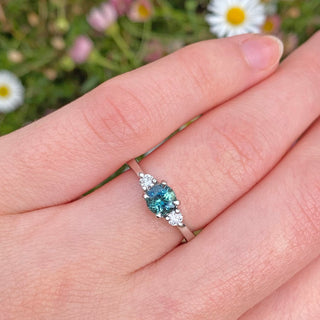 Blue teal sapphire and diamond trilogy engagement ring handmade from sustainable solid 950 platinum. Being worn on the hand, with daisy backdrop